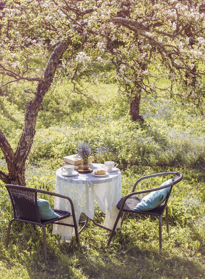 Springtime in garden. Table set with tea cups  boho reuse jar vase with forget me not flowers Myosotis and small stack of books. Summer relax readings concept. Springtime in garden. Table set with tea cups  boho reuse jar vase with forget me not flowers Myosotis and small stack of books. Summer relax readings concept.