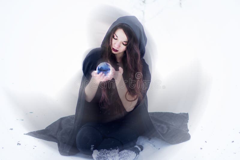 Witch or woman doing magic in black cloak with glass ball in white snow forest