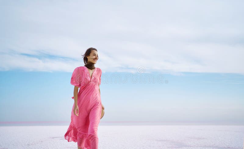 Freedom cheerfulness woman walking on white salty beach on pink salt lake with paradise view and beautiful blue sky. Elegant girl in stylish blowing by wind pink dress. Freedom cheerfulness woman walking on white salty beach on pink salt lake with paradise view and beautiful blue sky. Elegant girl in stylish blowing by wind pink dress