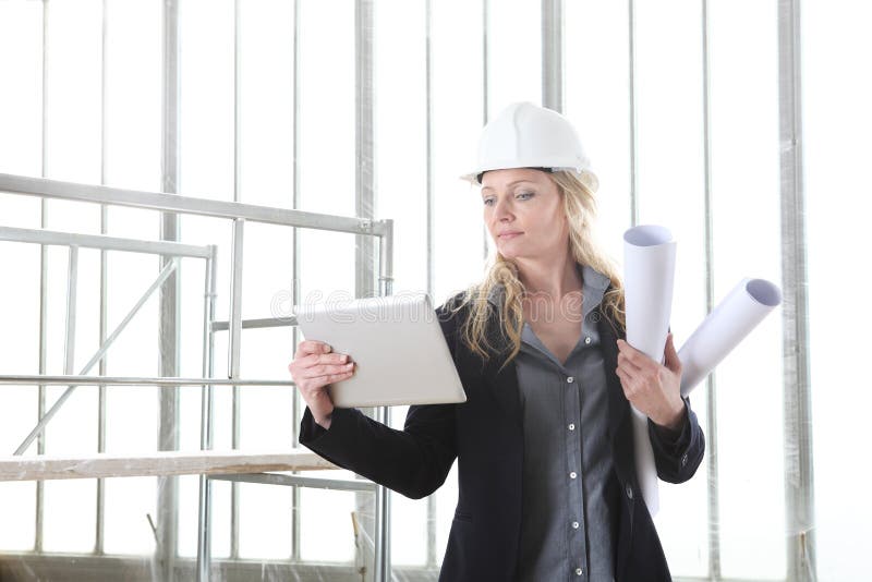 Woman architect or construction engineer with digital tablet wear helmet and holds blueprint inside a building site with windows royalty free stock photography