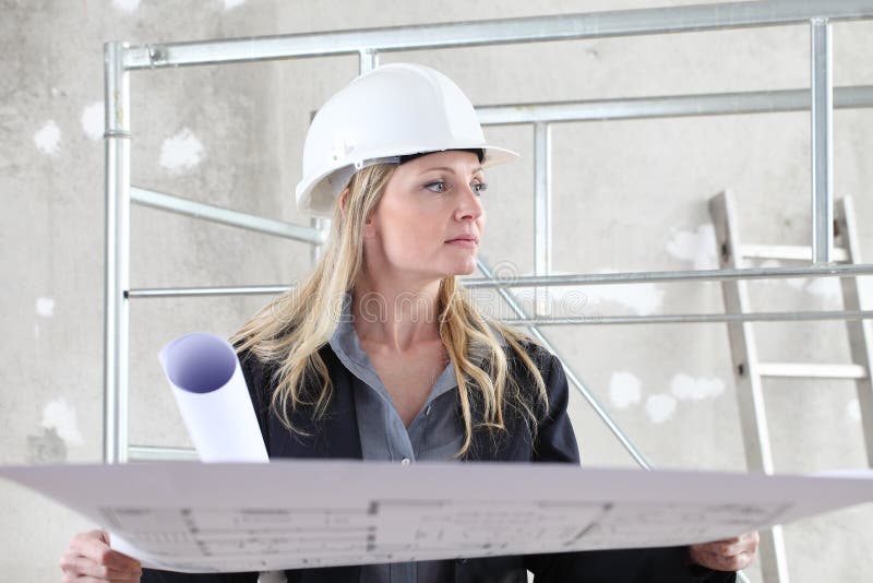 Woman architect or construction engineer reading blueprint wear helmet inside a building site with scaffolding in the background.