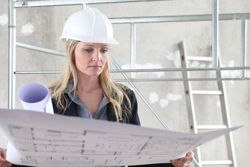 Woman architect or construction engineer reading blueprint wear helmet inside a building site with scaffolding in the background stock image