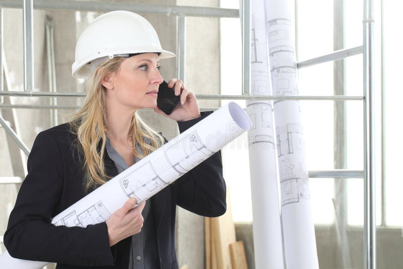 Woman architect or construction engineer talk on the mobile phone wear helmet and holds blueprint inside a building site with stock photos
