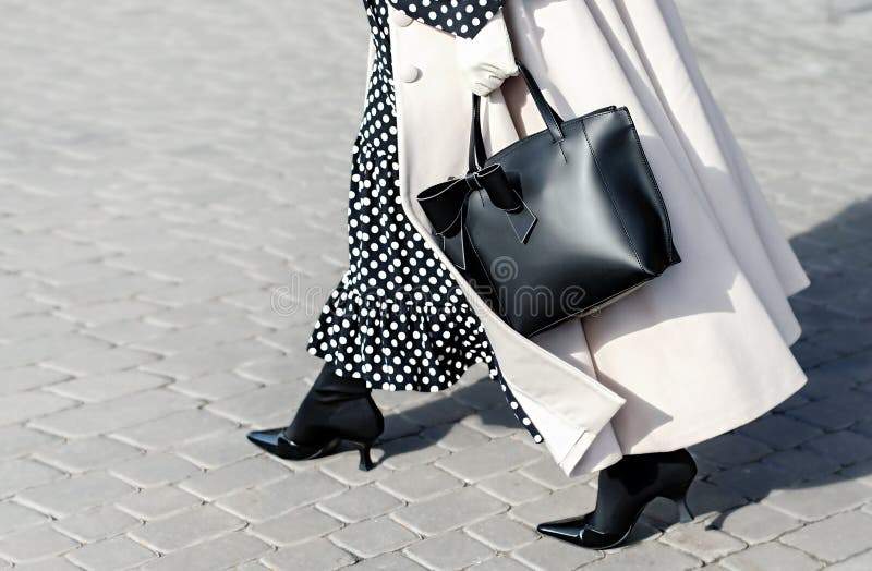 Woman in autumn or spring in a beige coat and a black dress with white polka dots. Fashionable bag close-up in female hands.