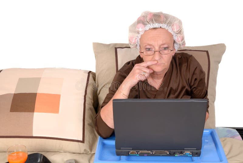Middle aged woman in pajamas working at home in bed, laptop on tray. Curlers and net in hair. Middle aged woman in pajamas working at home in bed, laptop on tray. Curlers and net in hair.