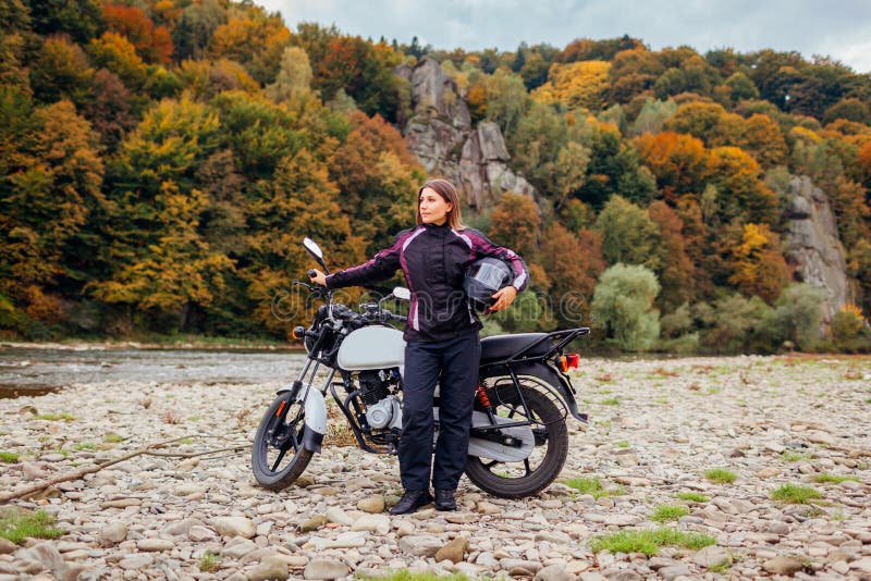 Woman biker travel by motorbike in fall. Motorcyclist enjoys autumn landscape in mountains having rest by forest