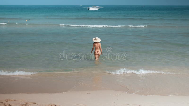 Woman in bikini and hat walks into ocean basking in radiant sun gentle caress of waves. Woman walks wades further