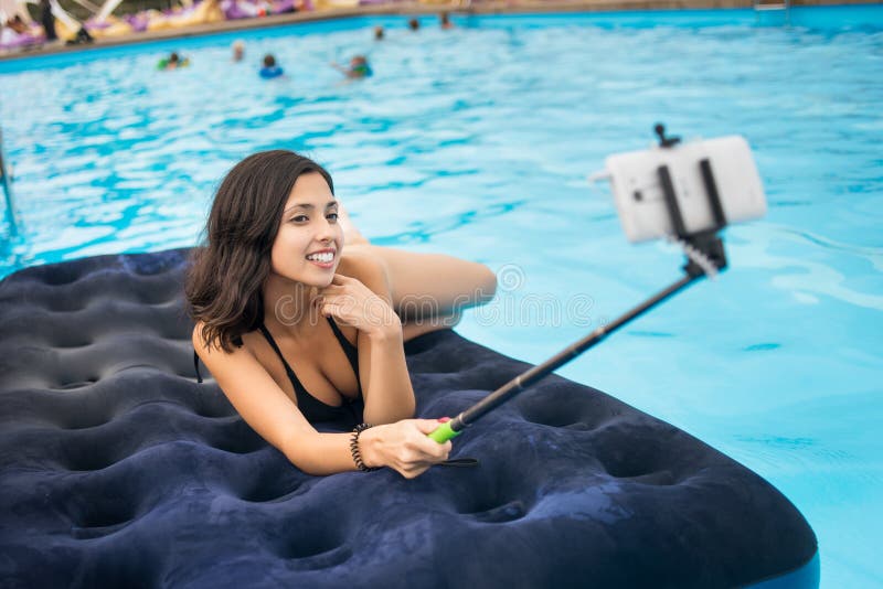 Attractive woman in bikini smiling and taking selfie photo on the phone with selfie stick on a mattress in the pool at the resort.