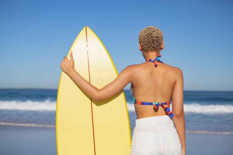 Rear view of African american woman in bikini standing with surfboard on beach in the sunshine