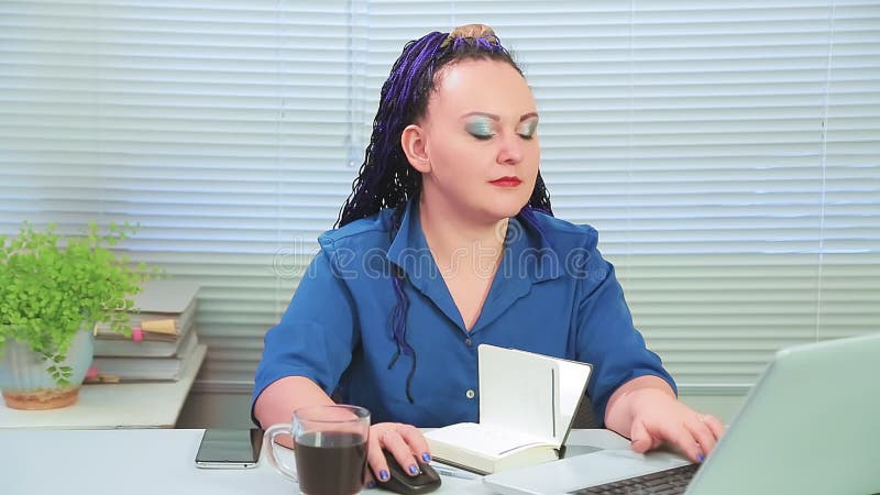 Woman with blue afro braids in the office with her back to the window at the computer drinks coffee