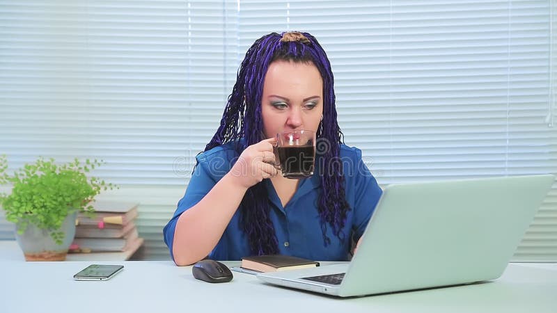 Woman with blue afro braids in the office with her back to the window at the computer drinks coffee