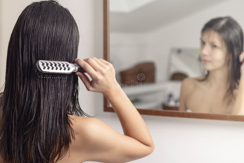 Young woman standing looking at her reflection in the bathroom mirror while brushing her wet hair with a hairbrush after washing it, view from behind. Young woman standing looking at her reflection in the bathroom mirror while brushing her wet hair with a hairbrush after washing it, view from behind