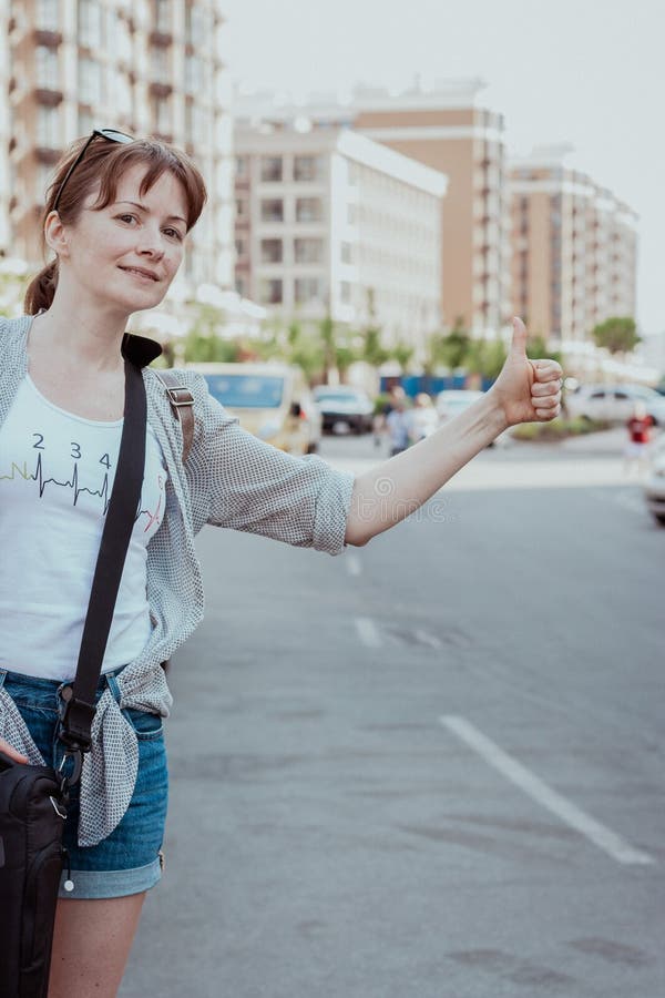 Woman catching a car. Stop car gesture. Journey by car. Girl waiting for taxi. Woman on a highway. Adventure concept. Smiling traveler on the street. Summer trip. Passenger waiting for auto.