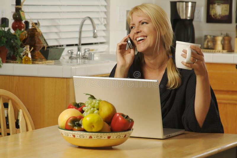 Woman smiling in her kitchen on cell phone sitting in front of laptop. Woman smiling in her kitchen on cell phone sitting in front of laptop.