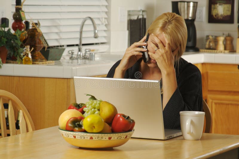 Woman holding her head in her kitchen on cell phone sitting in front of laptop. Woman holding her head in her kitchen on cell phone sitting in front of laptop.