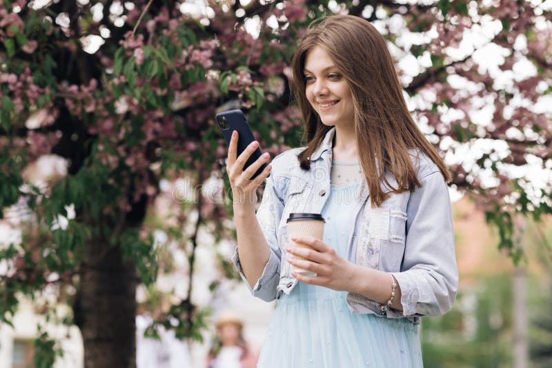 Woman with charming Smile walking in the Park and Using Mobile Phone Wearing Stylish Outfit. Girl Using Smartphone and