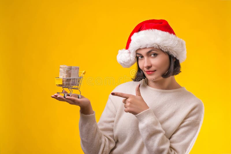 Woman in Christmas hat goes to shopping. Cute girl holding shopping cart with money, isolated on yellow. Woman thinking about royalty free stock photos Woman in Christmas hat goes to shopping. Cute girl holding shopping cart with money, isolated on yellow. Woman thinking about royalty free stock photos