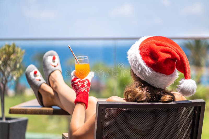 Woman in christmas santa claus hat and slippers relaxing and drinking cocktail on balсony opposite palm trees beach and sea.