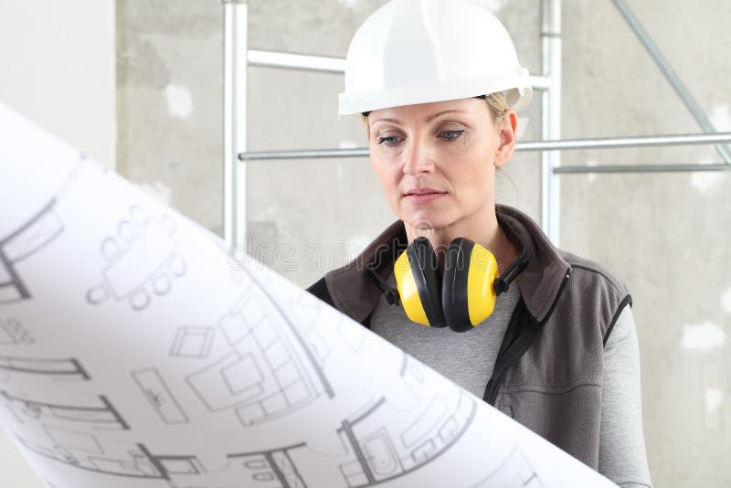 Woman construction worker builder looking at bluprint, wearing helmet and hearing protection headphones in building site indoors stock images