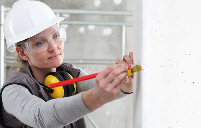 Woman construction worker work with meter tape and pencil, measure wall in interior building site, wearing helmet, glasses and royalty free stock images