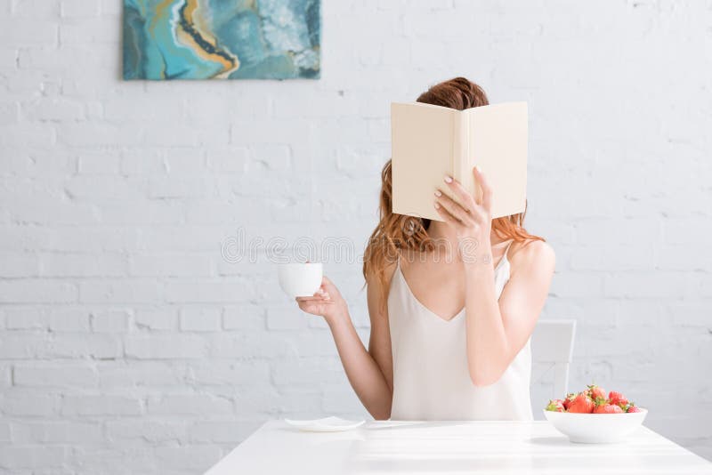 woman with cup of coffee and bowl of strawberries covering face with book