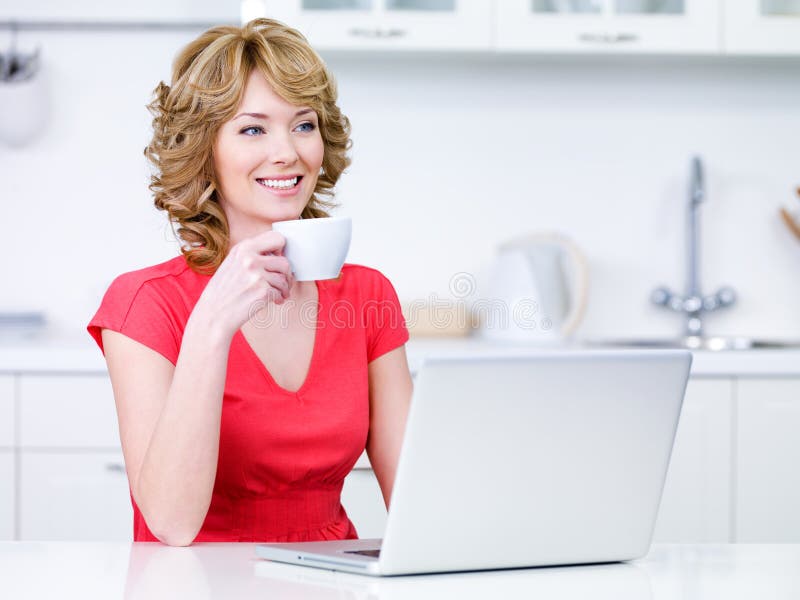 Young woman in casuals with cup of coffee and laptop in the kitchen. Young woman in casuals with cup of coffee and laptop in the kitchen