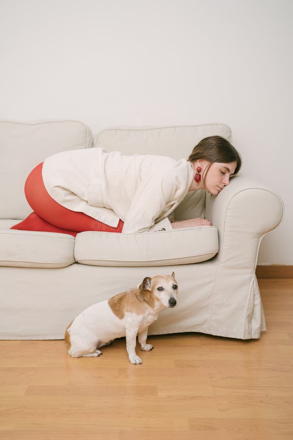 Woman curled up on beige sofa and dog on floor looking at camera.