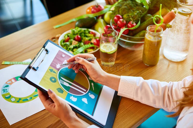 Woman dietitian in medical uniform with tape measure working on a diet plan sitting with different healthy food ingredients in the