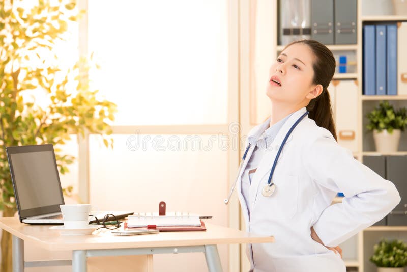 Attractive woman doctor with back pain in the clinic at hospital. mixed race asian chinese model. medical and healthcare concept.
