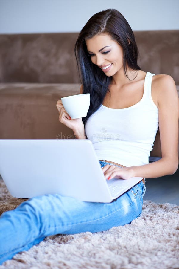 Woman drinking coffee as she types on her laptop computer while relaxing sitting on the carpet leaning against a sofa