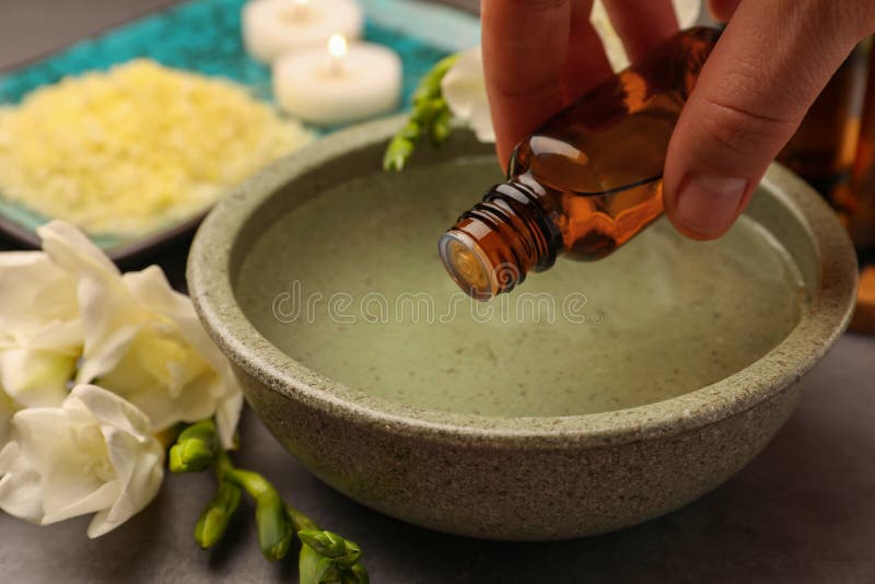 Woman dripping essential oil into bowl at grey table, closeup. Aromatherapy treatment