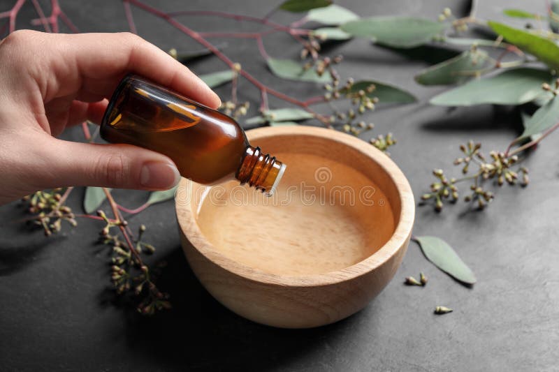 Woman dripping eucalyptus essential oil from bottle into bowl at grey table, closeup