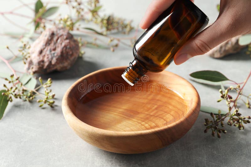Woman dripping eucalyptus essential oil from bottle into bowl at light grey table, closeup