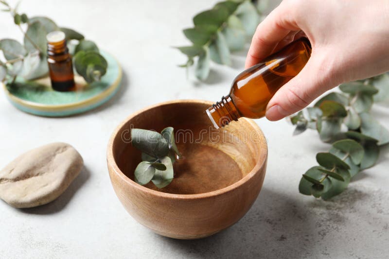 Woman dripping eucalyptus essential oil from bottle into bowl at light grey table, closeup