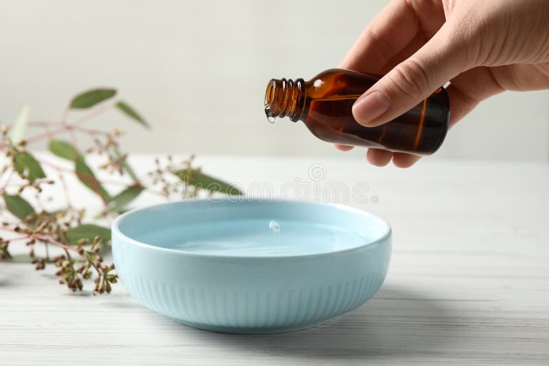 Woman dripping eucalyptus essential oil from bottle into bowl at white wooden table, closeup