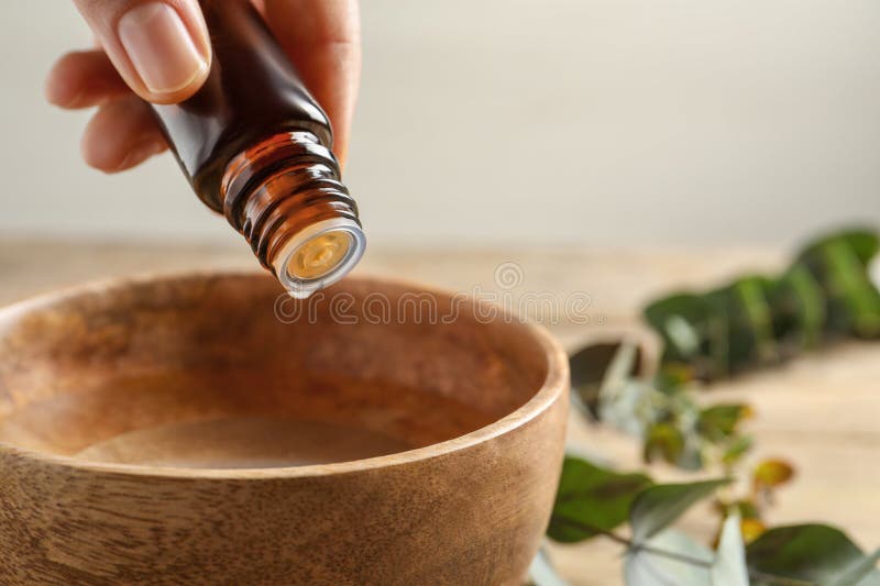 Woman dripping eucalyptus essential oil from bottle into bowl at wooden table, closeup. Space for text