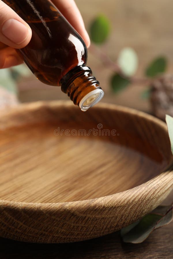 Woman dripping eucalyptus essential oil from bottle into bowl at wooden table, closeup