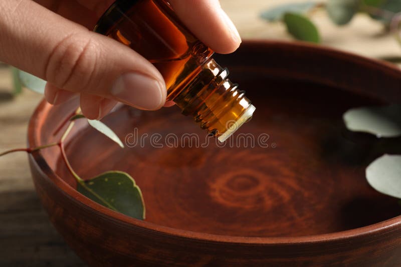 Woman dripping eucalyptus essential oil from bottle into bowl at wooden table, closeup