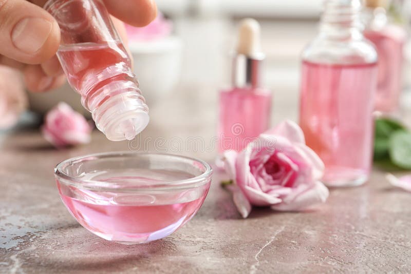 Woman dripping rose essential oil into bowl on table, closeup. Space for