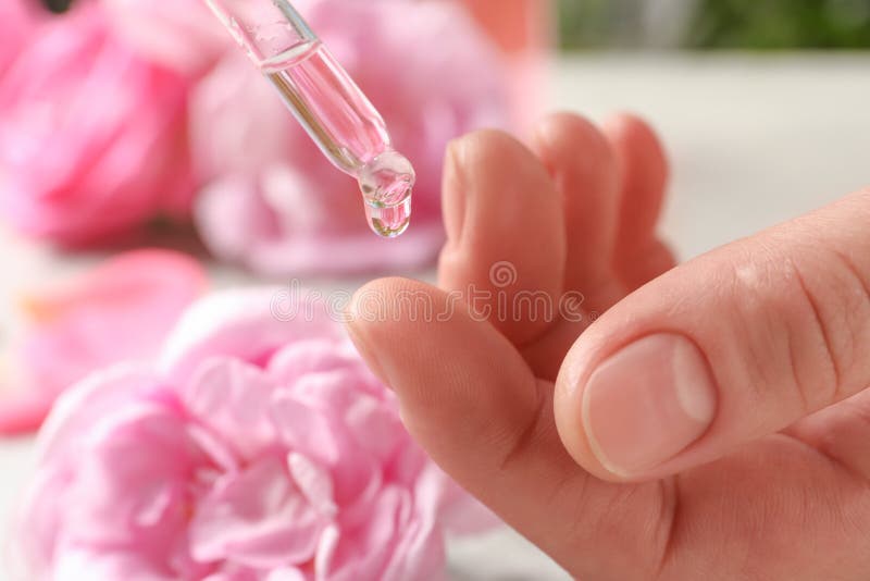 Woman dripping rose essential oil on finger against blurred flowers