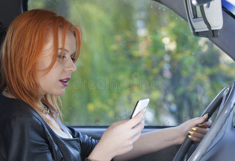 Concept of danger driving. Young woman driver red haired teenage girl texting on cell phone sending text reading message while driving the car. Concept of danger driving. Young woman driver red haired teenage girl texting on cell phone sending text reading message while driving the car.