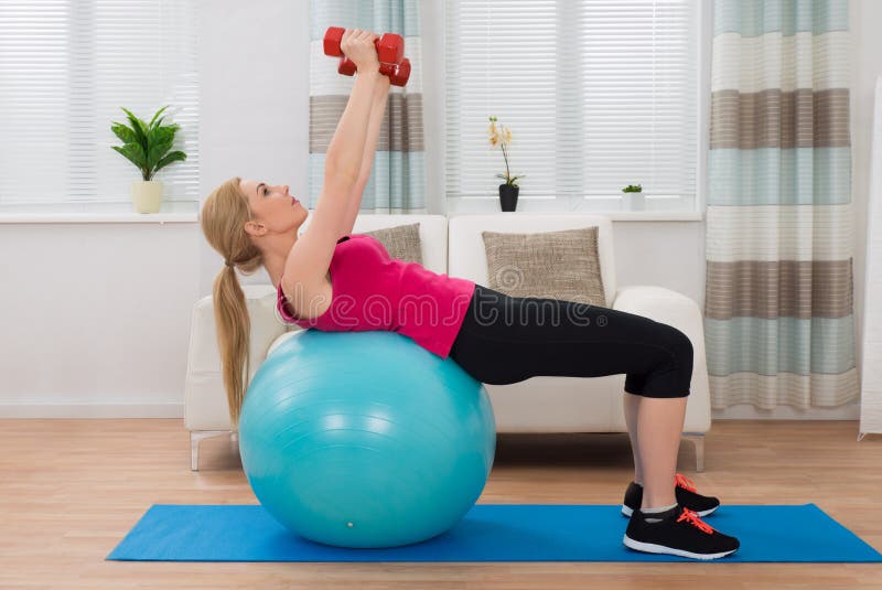 Woman With Dumbbell While Exercising On Fitness Ball. Young Woman Holding Dumbbell While Exercising On Fitness Ball In Living Room stock photo