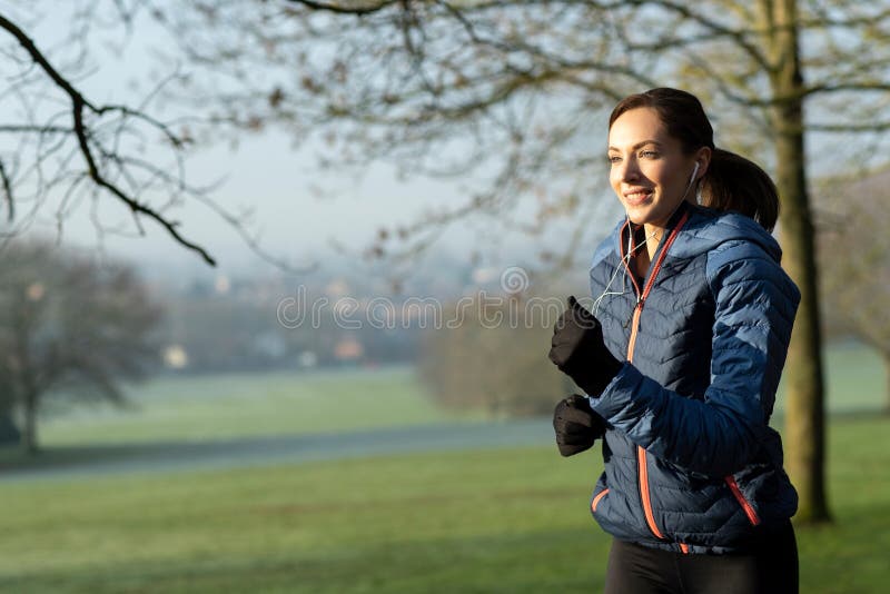 Woman On Early Morning Winter Run In Park Keeping Fit Listening To Music Through Earphones