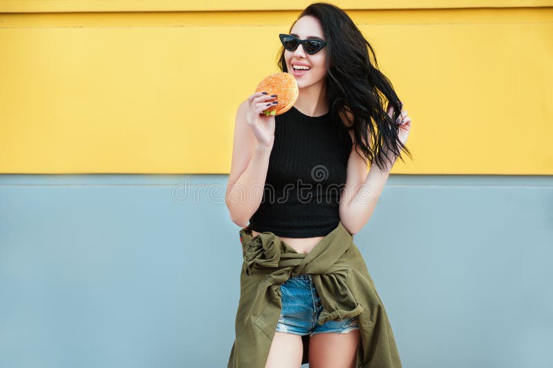 Woman eat Fast food. Portrait of beautiful girl in fashion casual clothes and trendy sunglasses laughing and eat burger outdoor on
