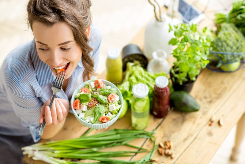 Woman eating healthy salad. Young and happy woman eating healthy salad sitting on the table with green fresh ingredients indoors royalty free stock photos
