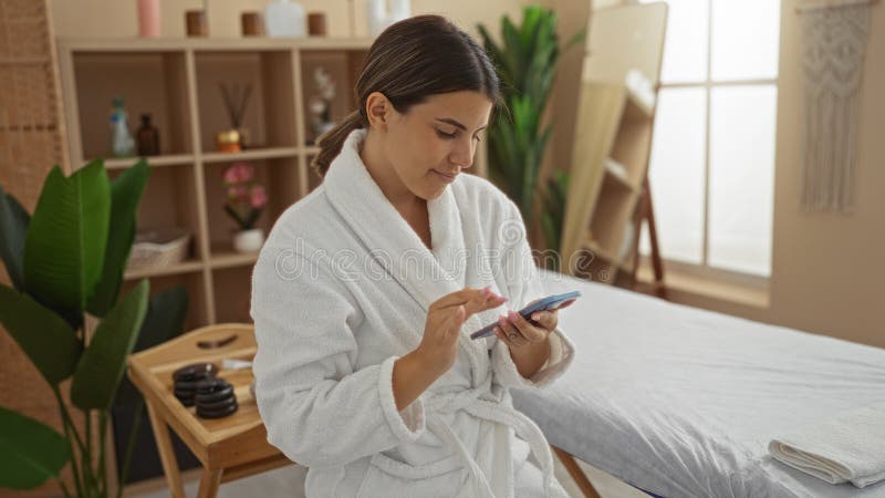 Woman enjoying a spa experience while using her smartphone in a tranquil wellness center with plants and massage table. Pampering experience stock images, royalty-free photos and pictures