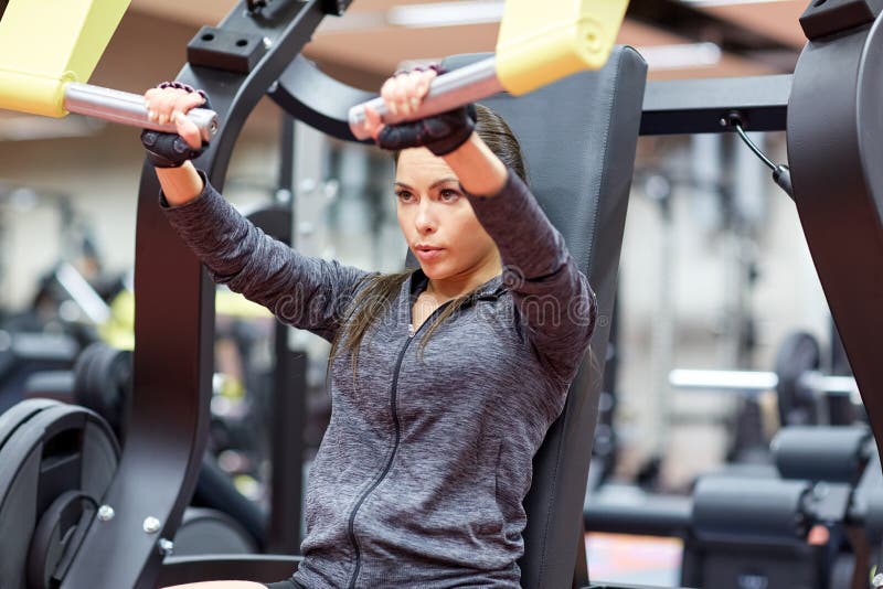 Fitness, sport, bodybuilding, exercising and people concept - young woman flexing muscles on seated chest press machine in gym. Fitness, sport, bodybuilding, exercising and people concept - young woman flexing muscles on seated chest press machine in gym