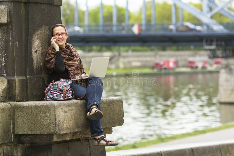 Woman freelancer sitting on the stone embankment of the river with a laptop and talking on the phone