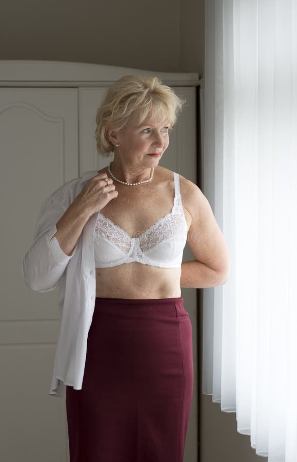 Elderly woman getting dressed putting on a shirt over a white bra standing ...