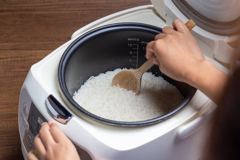 Woman hand is scooping jasmine rice cooking in electric rice cooker with steam. Thai Jasmine rice stock photo